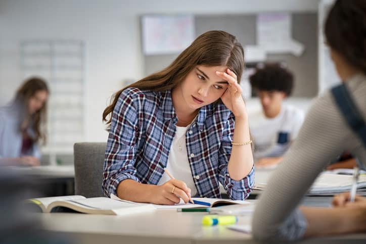 Anxious young woman with hand on head feeling tired while studying at school in classroom. College student suffering from headache in library preparing for test. Portrait of troubled and stressed girl doing exam that doesn"u2019t know the answers.