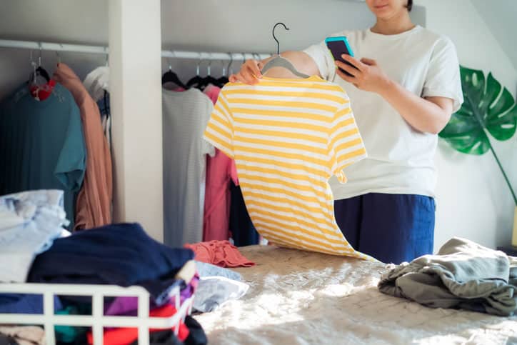 Woman taking photo of t-shirt using her smartphone for selling or donating her clothes. Decluttering Clothes, Sorting, And Cleaning Up. Reuse, second-hand concept. Conscious consumer.