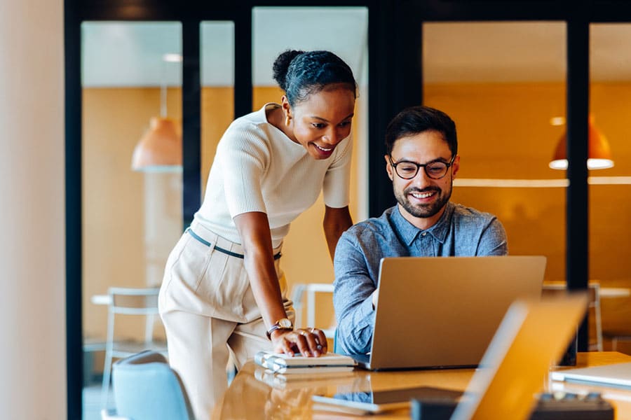 A woman and a man work together at a desk in a modern office. The hard worker is seated, smiling at his laptop, while the woman stands beside him, leaning in and looking at the screen.