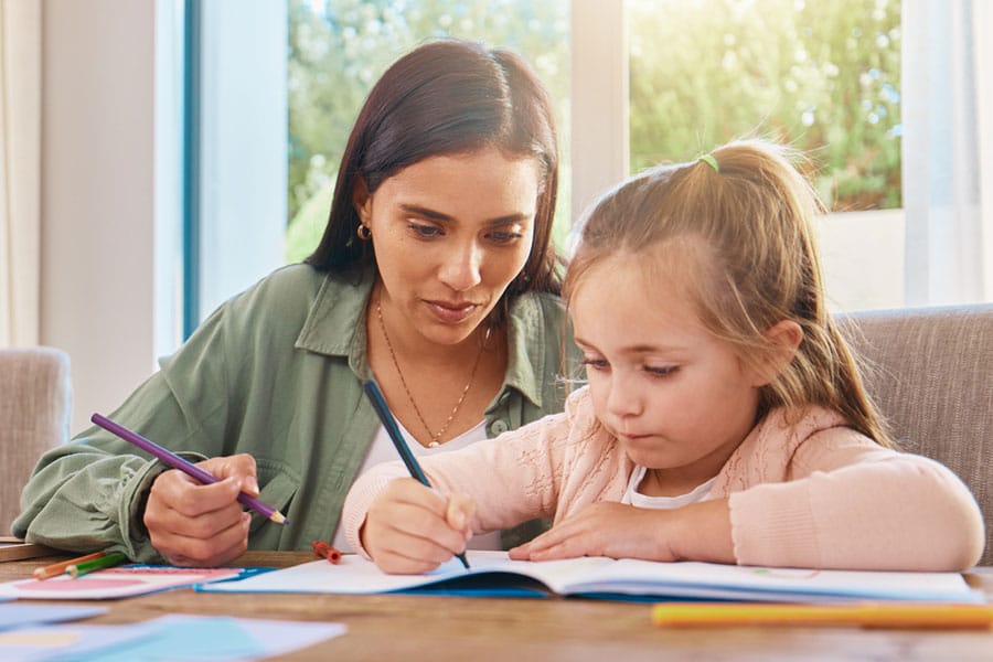 An adult and a young girl sit at a table, coloring or drawing together in a bright room with large windows and greenery outside, highlighting the importance of classroom support in fostering creativity and growth.