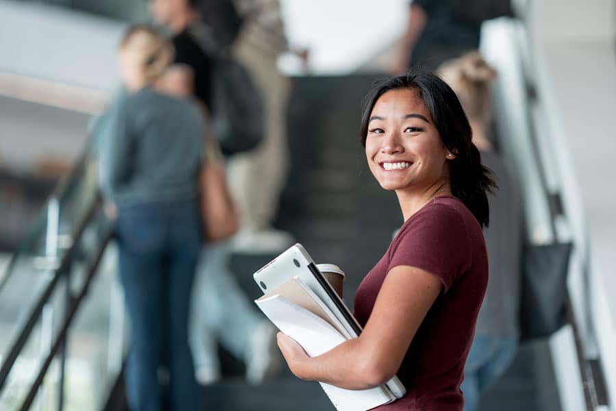 A young woman holding notebooks and a coffee cup smiles at the camera while standing near an escalator with several college students in the background.