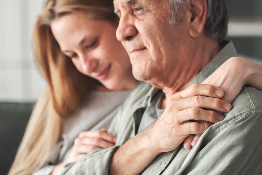 A young woman with a gentle expression rests her hand on the shoulder of an older man undergoing a cognitive evaluation.